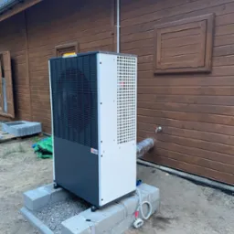 Zewnętrzna jednostka pompy ciepła, zamontowana na betonowej podstawie przy drewnianym budynku z brązową elewacją, widoczny przewód odprowadzający skropliny.