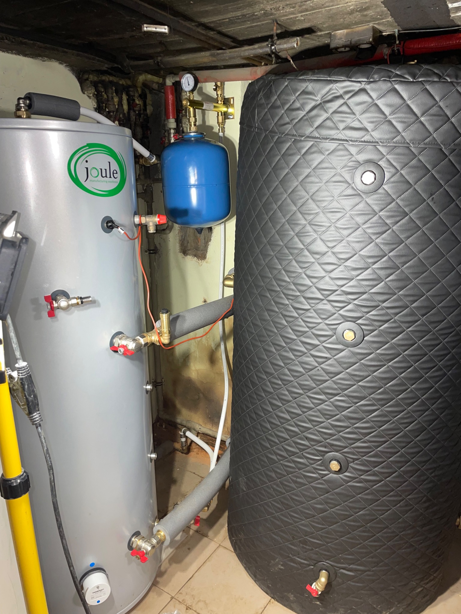 Dwa zbiorniki akumulacyjne ciepła: szary z logo Joule i czarny z pikowaną izolacją, połączone instalacją hydrauliczną w pomieszczeniu technicznym.