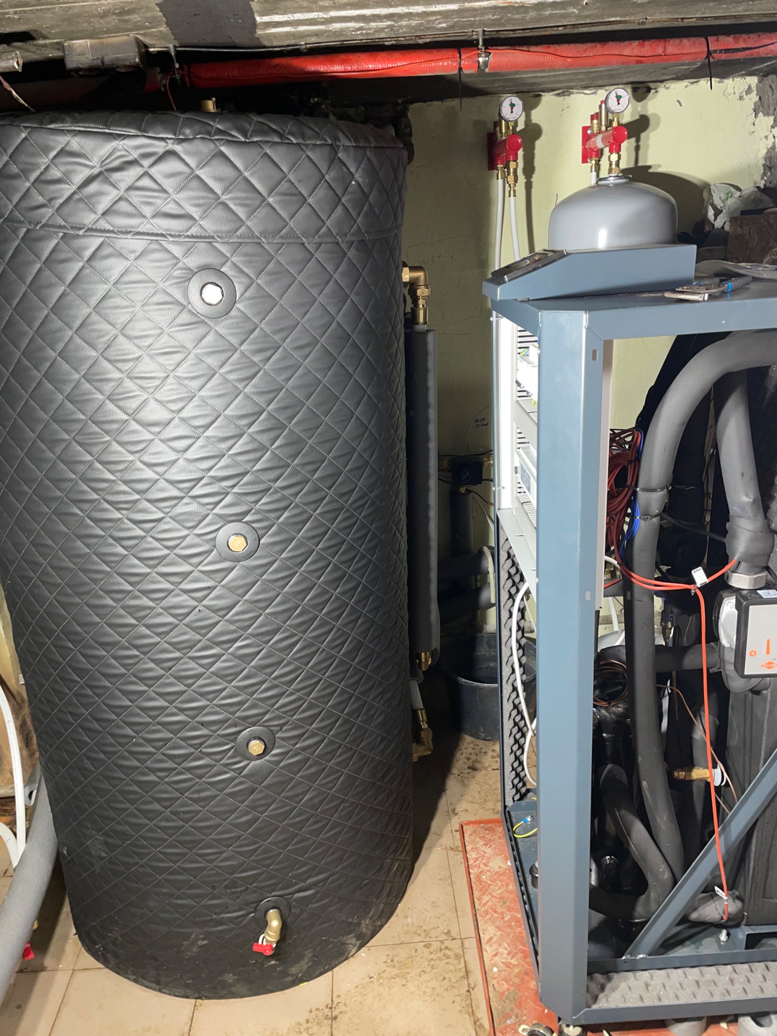 Duży, czarny, izolowany zbiornik akumulacyjny z widocznymi przyłączami, obok jednostka zewnętrzna z rurami i przewodami w szarej metalowej obudowie. Manometry na ścianie. Zdjęcie zrobione w piwnicy.