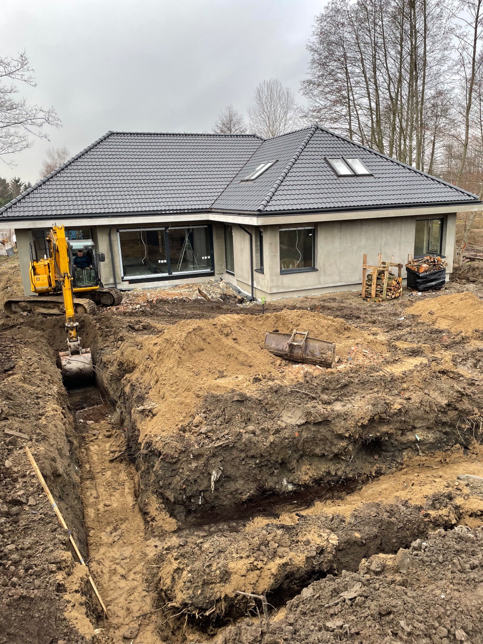 Żółta koparka wykopuje wąski rów pod fundamenty domu jednorodzinnego z szarą dachówką, widoczne palety z drewnem i stos czarnej folii.