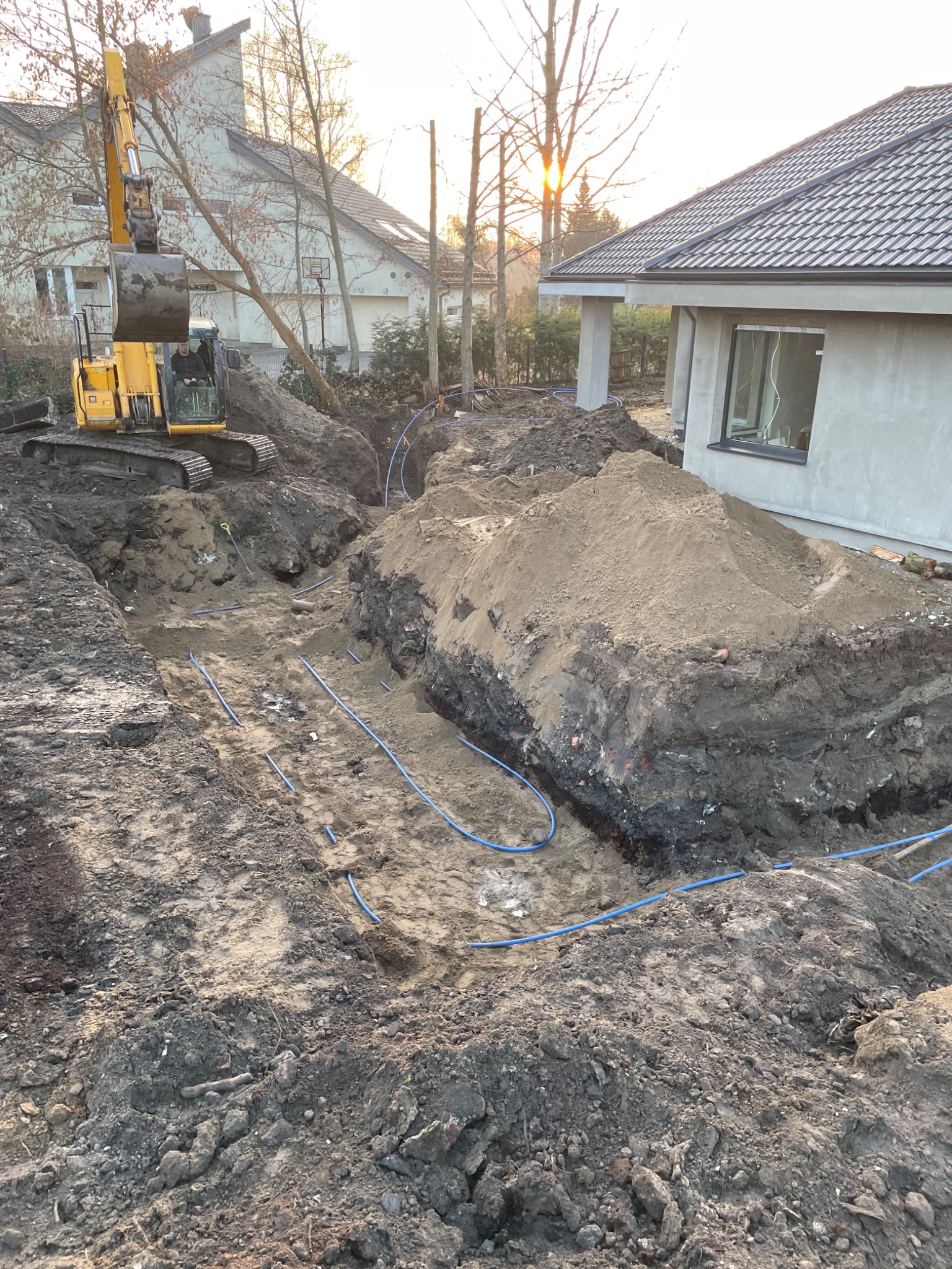 Żółta koparka pracująca przy wykopie z niebieskimi rurami, prawdopodobnie instalacja pompy ciepła w trakcie budowy domu jednorodzinnego.