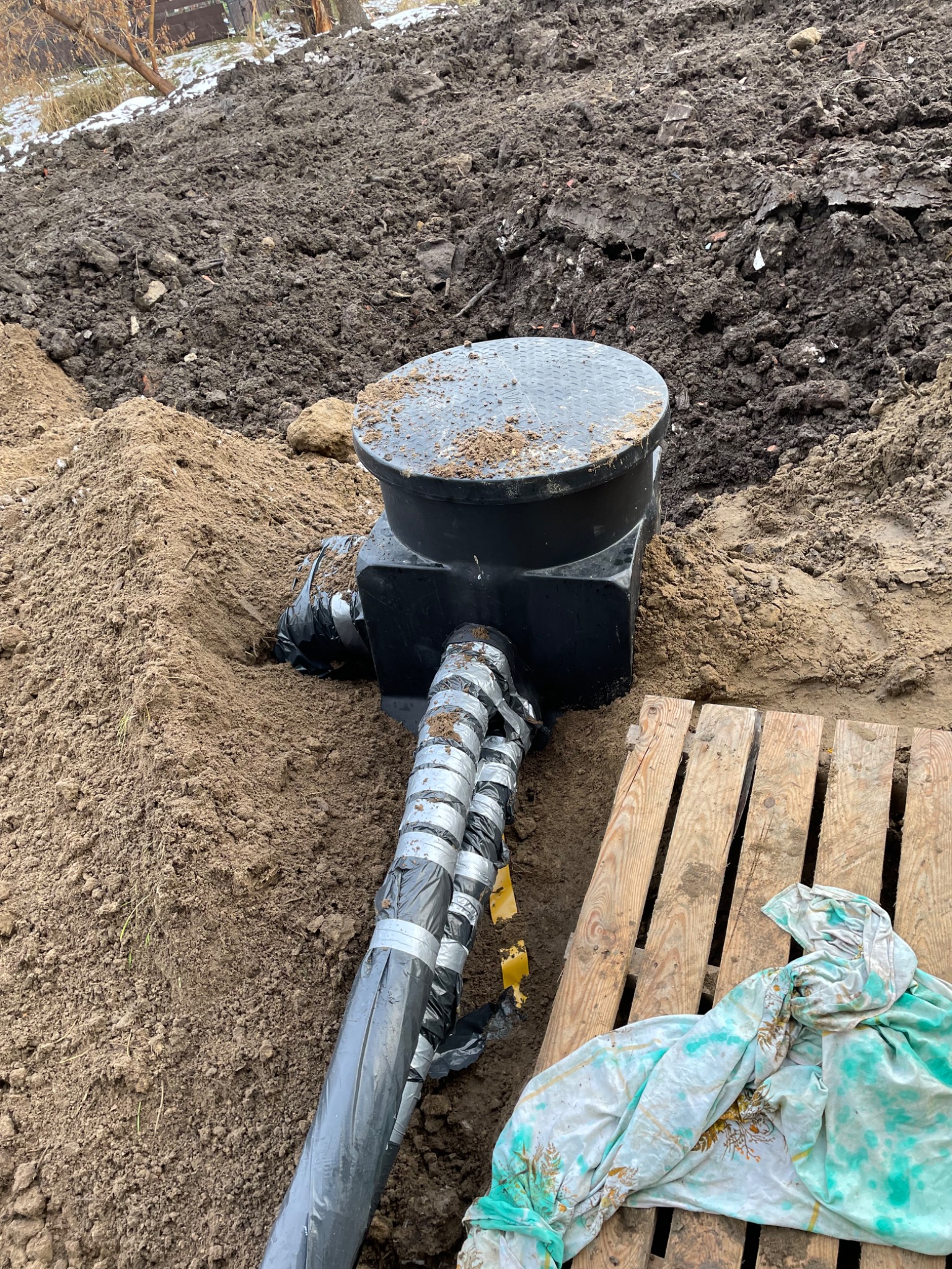 Czarna studzienka kanalizacyjna z rurami, zabezpieczonymi taśmą, w wykopie ziemnym, obok drewnianej palety przykrytej tkaniną.