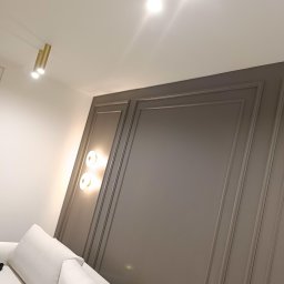 Szara ściana z dekoracyjnymi panelami i oświetleniem punktowym, widoczny fragment białej sofy. Nowoczesne wykończenie wnętrza z dbałością o detale.