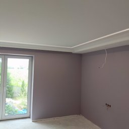 Pomieszczenie po malowaniu ścian na fioletowo, widoczne puszki z farbą, listwa dekoracyjna pod sufitem i wystające kable ze ściany. Okno z widokiem na zieleń.