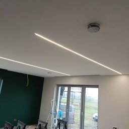 Nowoczesne oświetlenie liniowe LED w suficie podwieszanym, widoczne czarne kozły malarskie i drabina, ściana pomalowana na zielono. W tle okno.