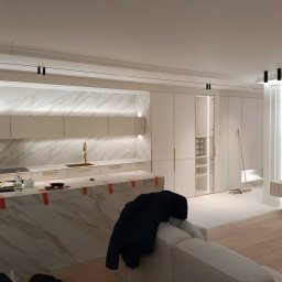 Dadex - Nowoczesna kuchnia z marmurowym blatem wyspowym zabezpieczonym taśmą, białe szafki z złotymi uchwytami i marmurowe ściany. Widoczne narzędzia i akcesoria na blacie.