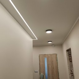 Dadex - Nowoczesne oświetlenie LED w korytarzu z ukrytą listwą świetlną i okrągłymi lampami sufitowymi. Widoczne przygotowania do montażu ościeżnicy drzwiowej.