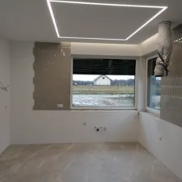 Wnętrze w trakcie wykańczania: szare płytki na podłodze, fragment ściany z dekoracyjnymi, podłużnymi płytkami, okno z widokiem na dom i pole, oraz nowoczesne oświetlenie sufitowe w kształcie kwadratu.