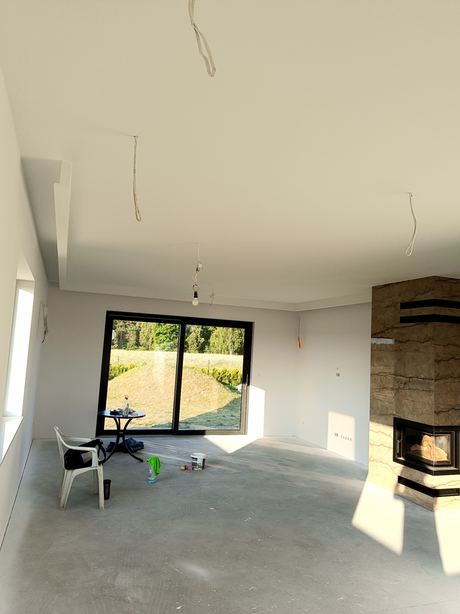 Jasne wnętrze po pracach wykończeniowych: gładkie ściany, sufit z listwami, marmurowy kominek. Widoczne kable oświetleniowe, okno z widokiem na zieleń i akcesoria malarskie.