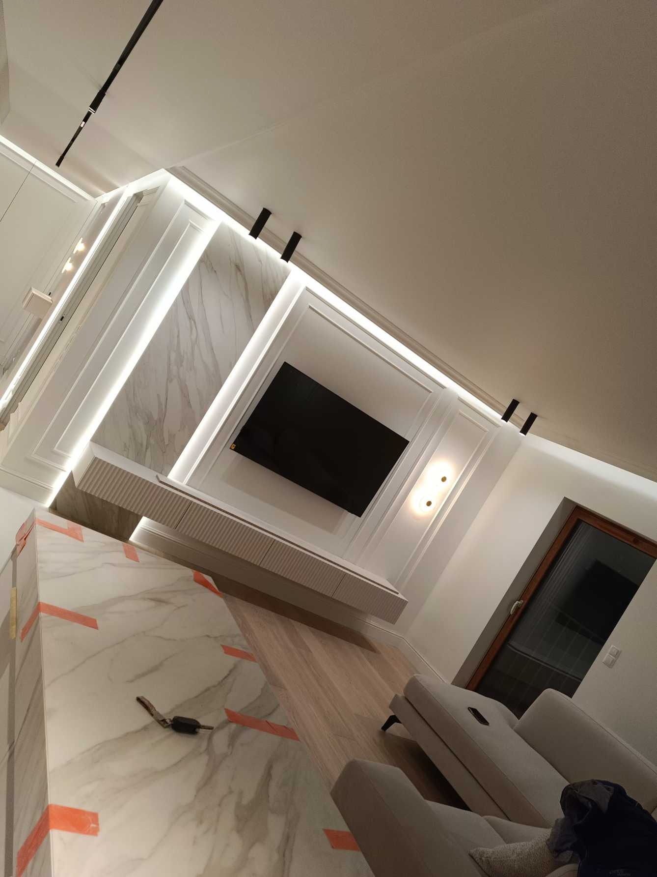 Nowoczesne wykończenie wnętrza salonu z marmurową ścianą, podświetleniem LED, drewnianą podłogą i minimalistycznym telewizorem. Widok na okno i część sofy.