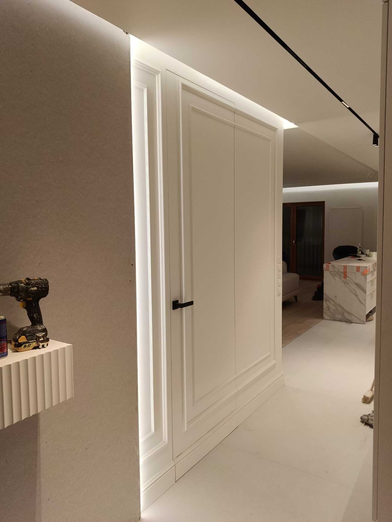 Białe, eleganckie drzwi z czarną klamką, wkomponowane w ścianę z oświetleniem LED. W tle widoczny fragment wnętrza z marmurowym blatem i oświetleniem punktowym.