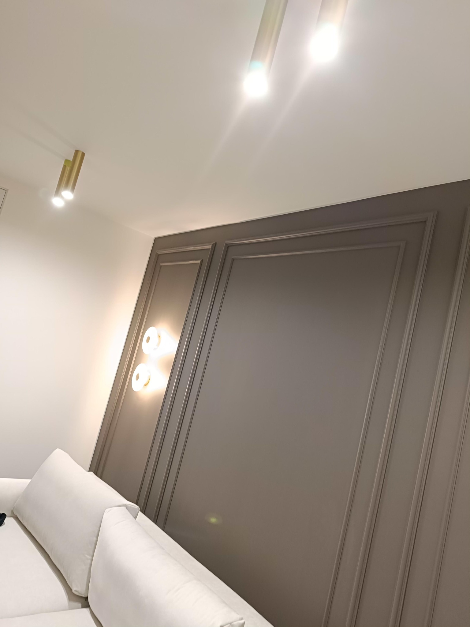 Szara ściana z dekoracyjnymi panelami i oświetleniem punktowym, widoczny fragment białej sofy. Nowoczesne wykończenie wnętrza z dbałością o detale.