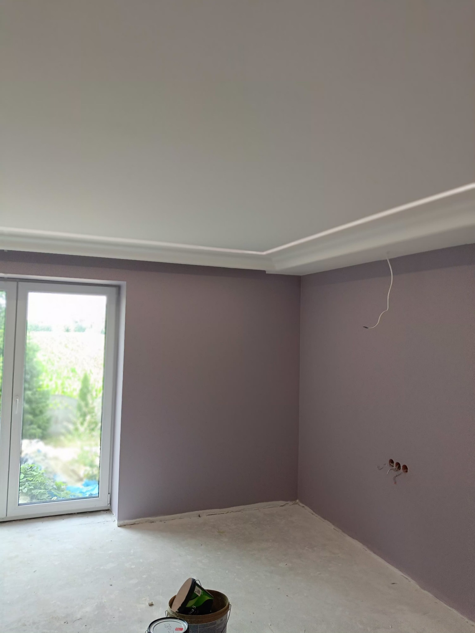 Pomieszczenie po malowaniu ścian na fioletowo, widoczne puszki z farbą, listwa dekoracyjna pod sufitem i wystające kable ze ściany. Okno z widokiem na zieleń.