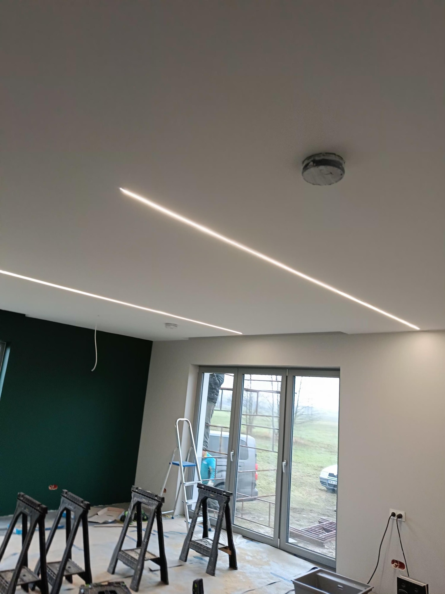 Nowoczesne oświetlenie liniowe LED w suficie podwieszanym, widoczne czarne kozły malarskie i drabina, ściana pomalowana na zielono. W tle okno.