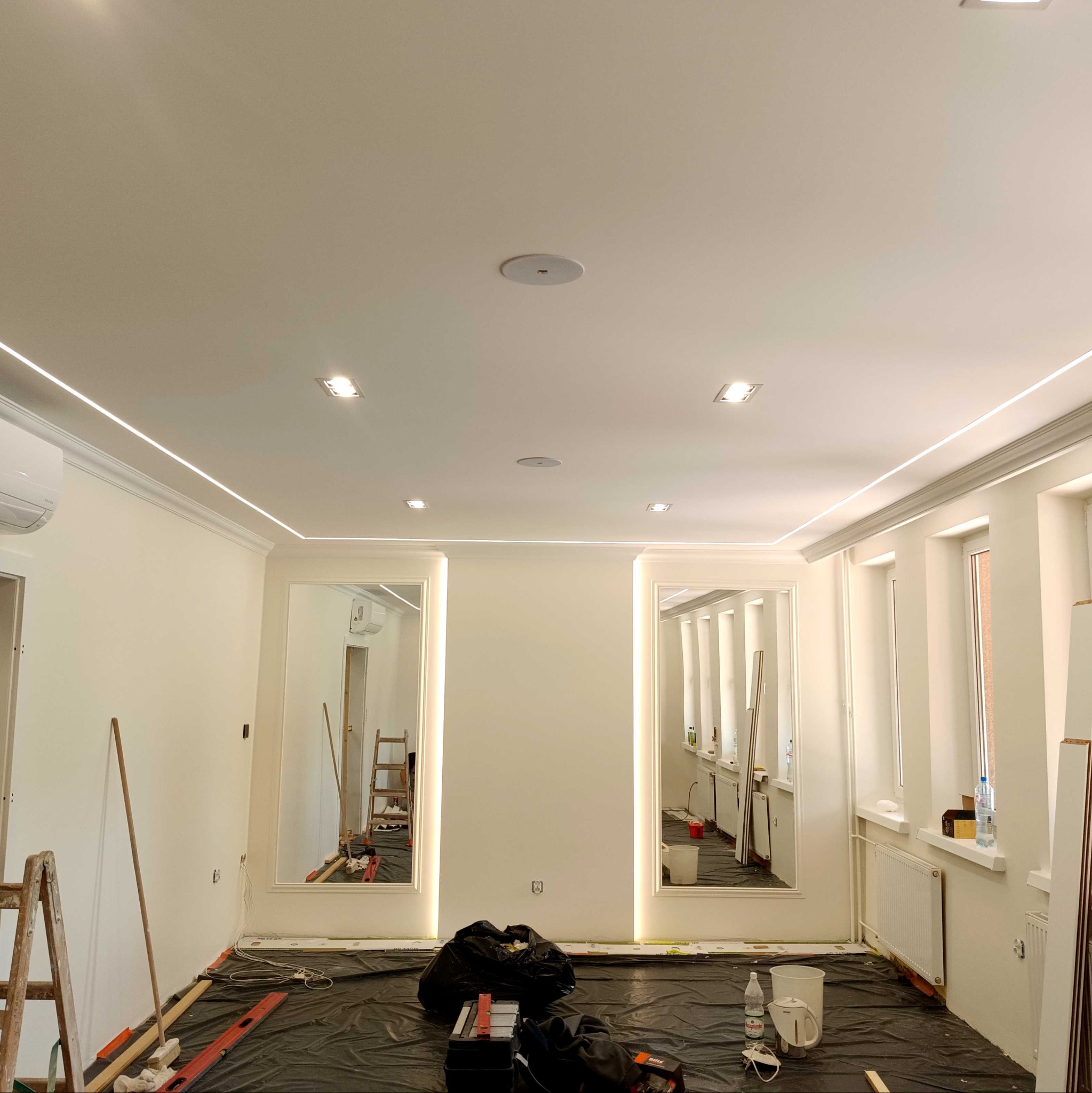 Jasne wnętrze w trakcie prac wykończeniowych: świeżo pomalowane ściany, listwy LED pod sufitem, zabezpieczona podłoga, drabina i narzędzia w tle. Widoczne lustra.