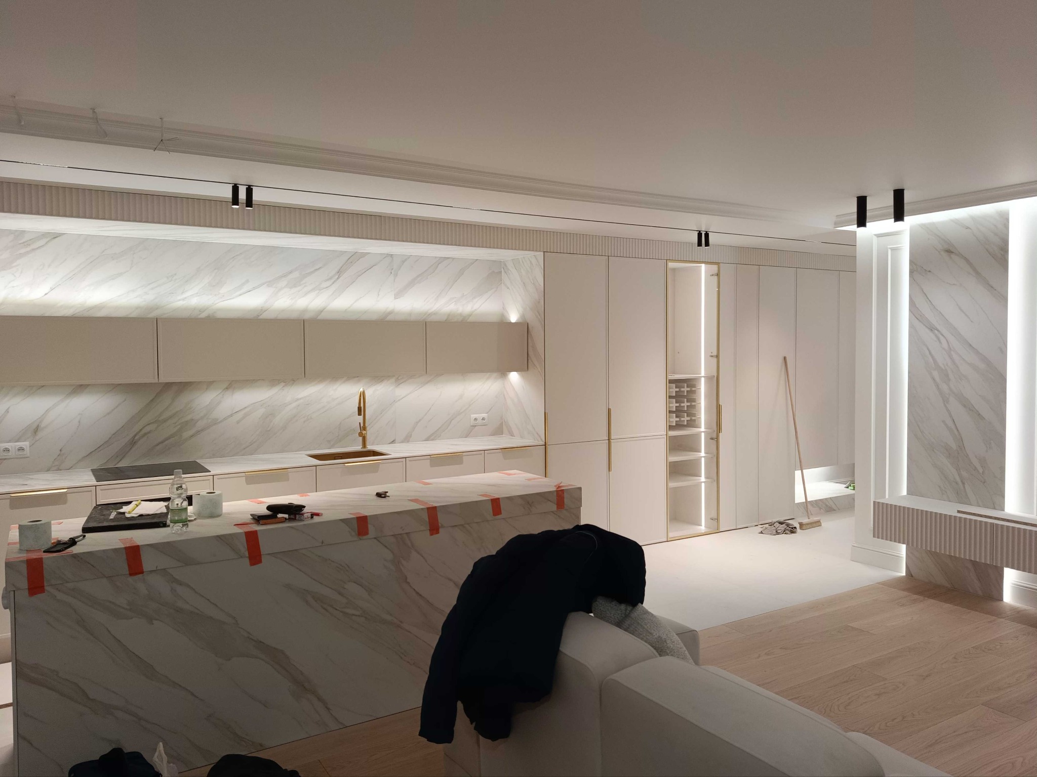 Nowoczesna kuchnia z marmurowym blatem wyspowym zabezpieczonym taśmą, białe szafki z złotymi uchwytami i marmurowe ściany. Widoczne narzędzia i akcesoria na blacie.