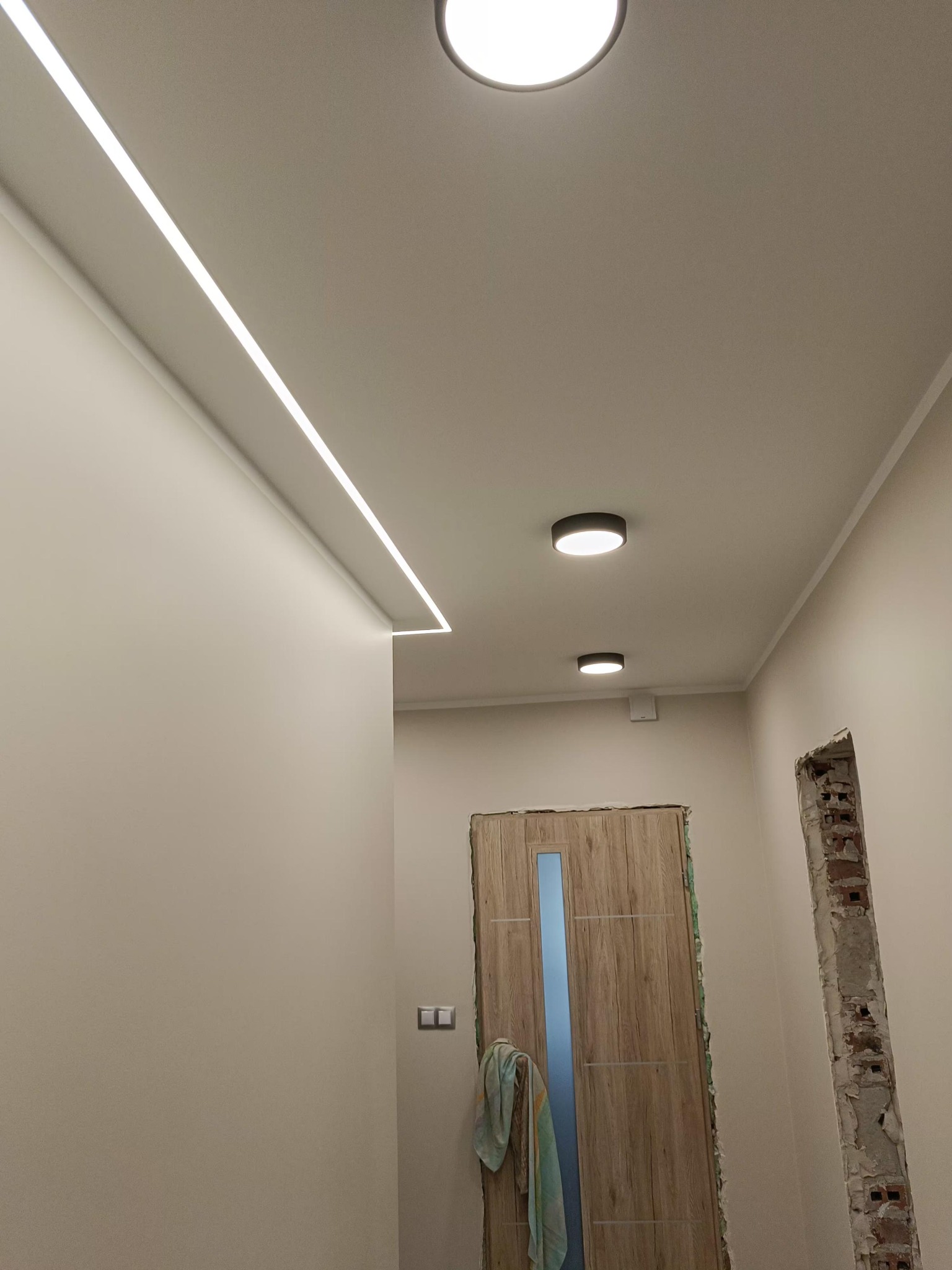 Nowoczesne oświetlenie LED w korytarzu z ukrytą listwą świetlną i okrągłymi lampami sufitowymi. Widoczne przygotowania do montażu ościeżnicy drzwiowej.