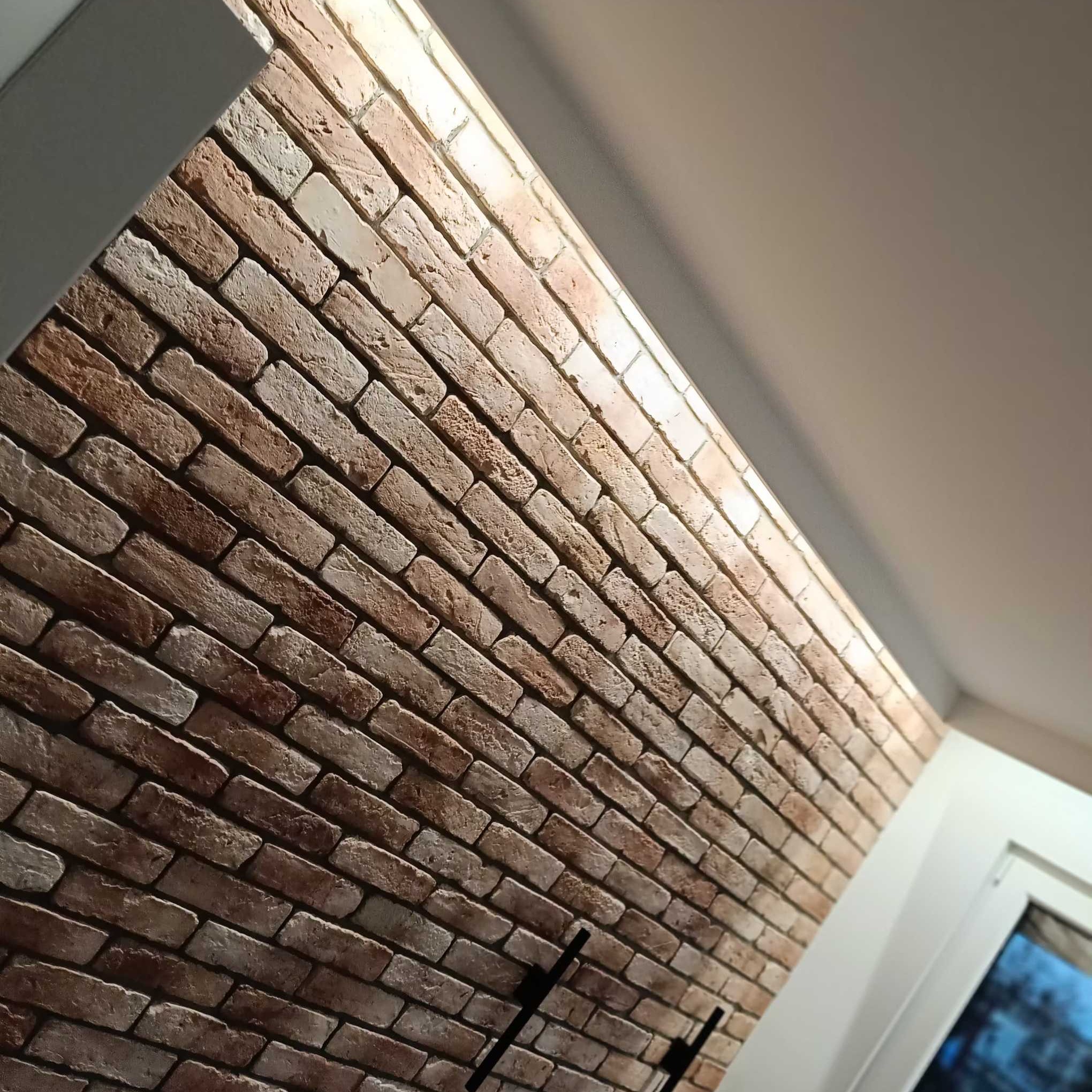 Ściana z cegieł dekoracyjnych w odcieniach brązu i beżu, oświetlona od góry, z fragmentem okna w tle. Układ cegieł pod kątem, widoczna faktura i nierówności.