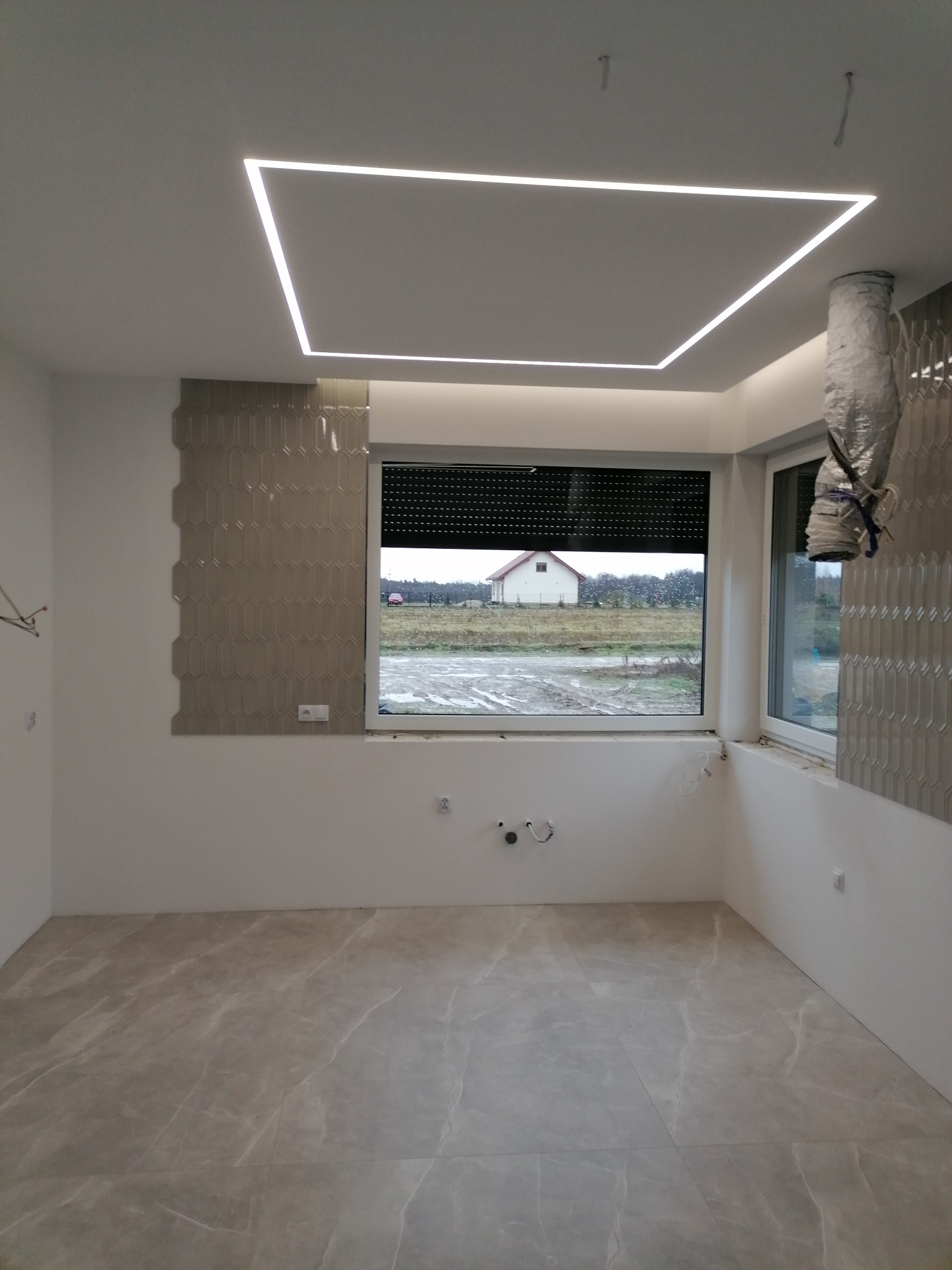 Wnętrze w trakcie wykańczania: szare płytki na podłodze, fragment ściany z dekoracyjnymi, podłużnymi płytkami, okno z widokiem na dom i pole, oraz nowoczesne oświetlenie sufitowe w kształcie kwadratu.