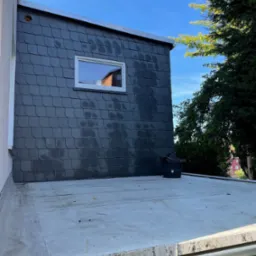 Elewacja budynku wykończona ciemnoszarą łupkową okładziną z małym oknem, widok z płaskiego dachu z widocznym koszem na śmieci i drzewami w tle.