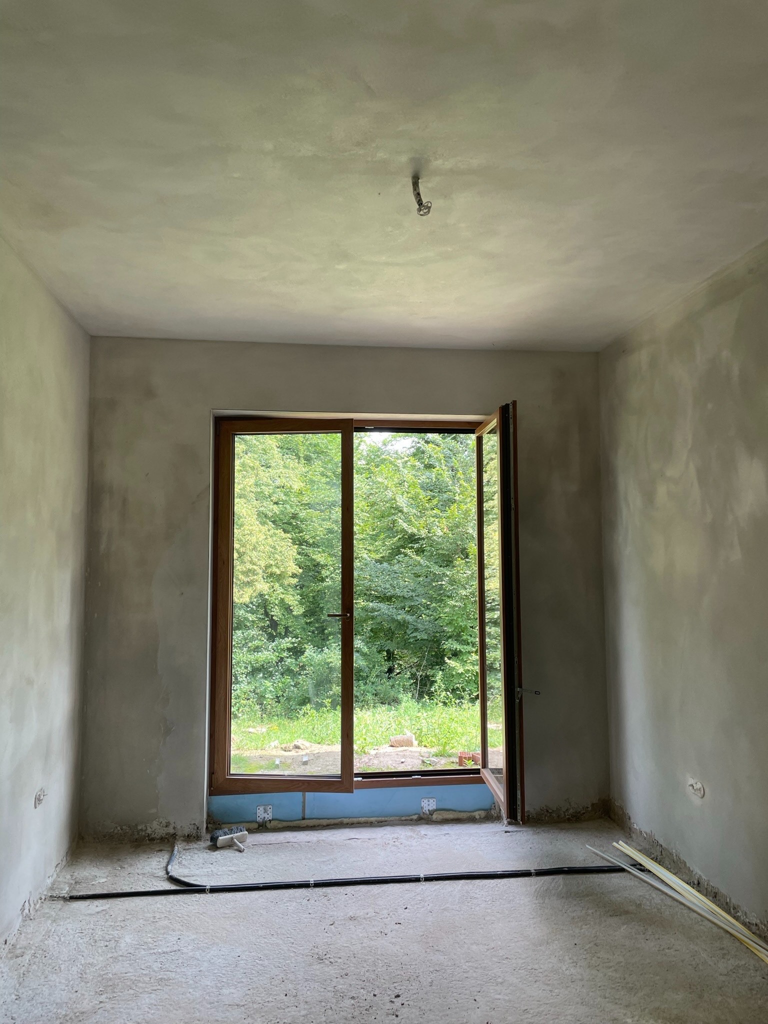 Pokój w trakcie remontu z gładzią na ścianach, otwartym oknem z widokiem na zieleń i widocznymi przewodami na podłodze.