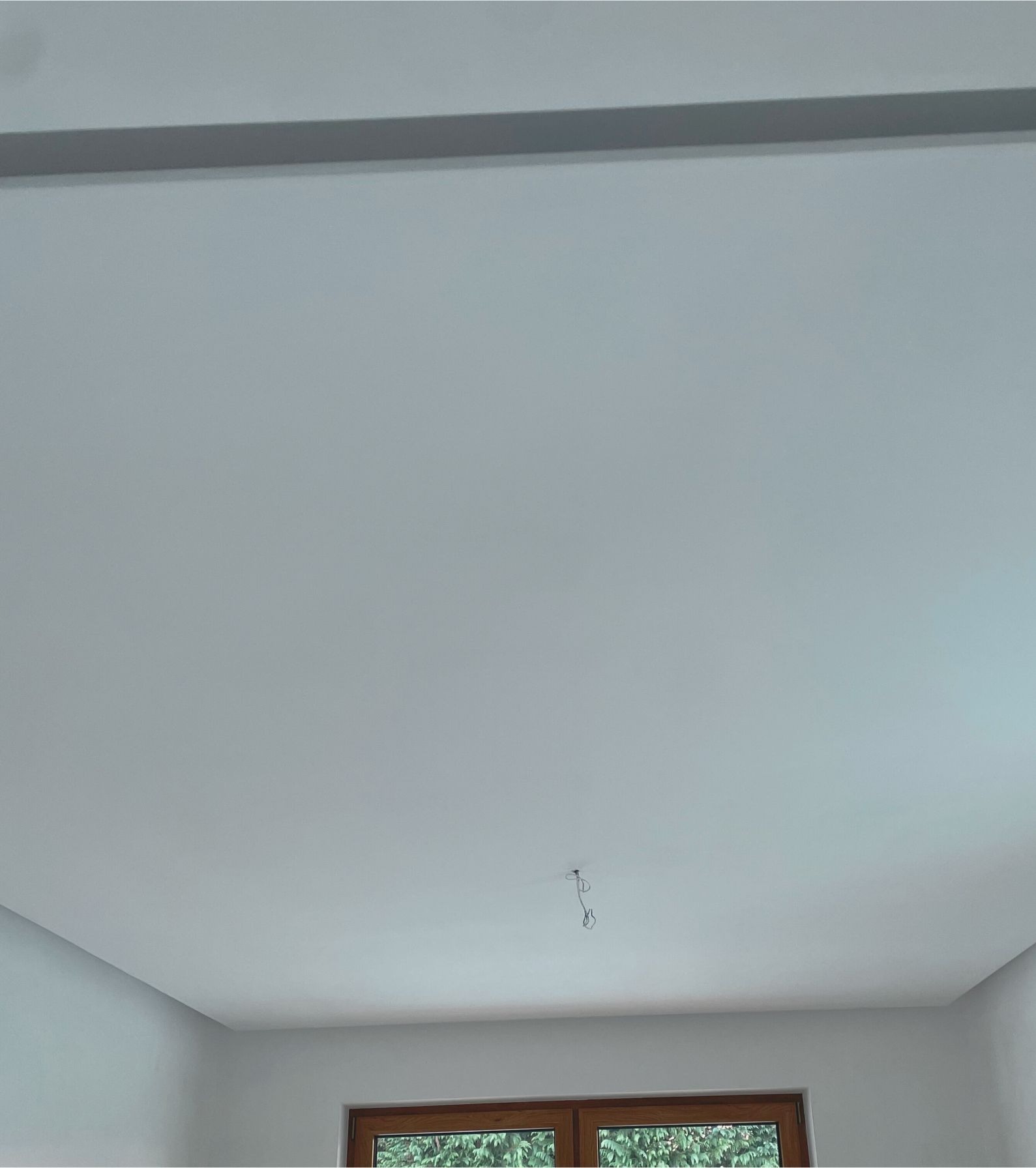 Gładki, biały sufit z widocznym kablem wystającym pośrodku, fragment ściany i okna z widokiem na zieleń na dole kadru.