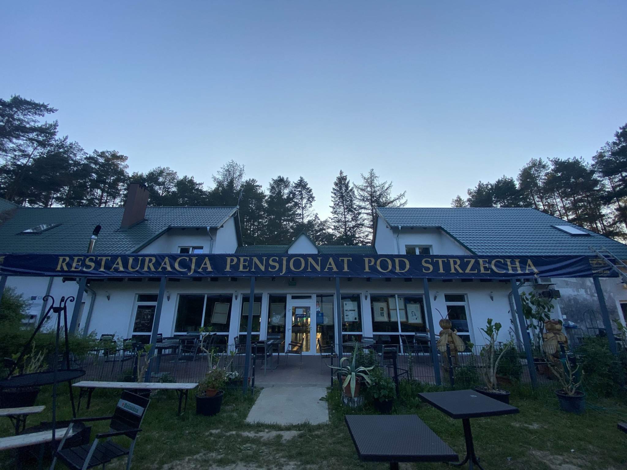 Fasada restauracji i pensjonatu 'Pod Strzecha' z widocznym banerem reklamowym, stolikami na zewnątrz i otaczającym lasem wczesnym rankiem.