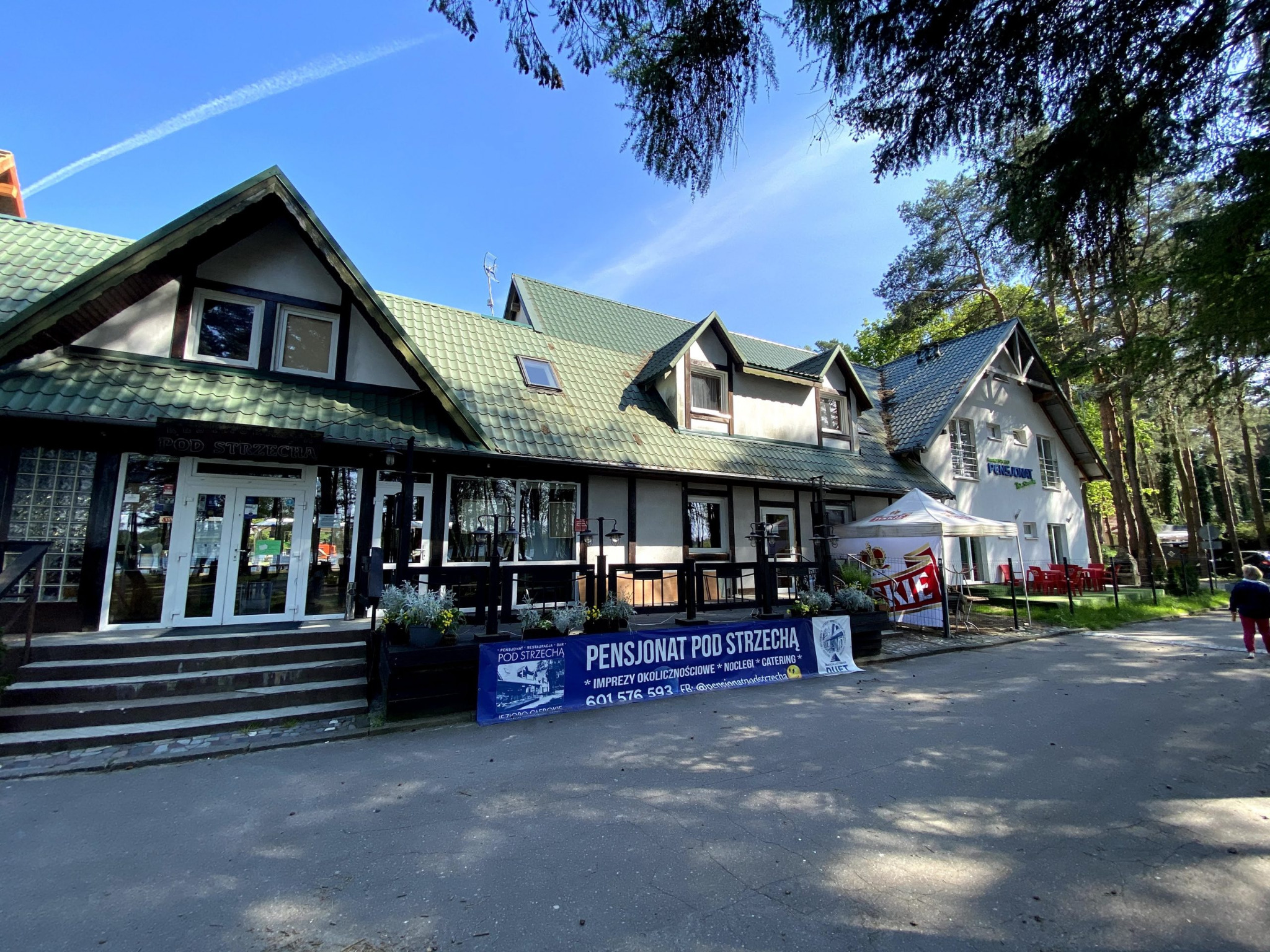 Pensjonat z zielonym dachem, widok z zewnątrz, baner reklamowy z ofertą noclegów i cateringu, błękitne niebo z śladem kondensacyjnym samolotu.