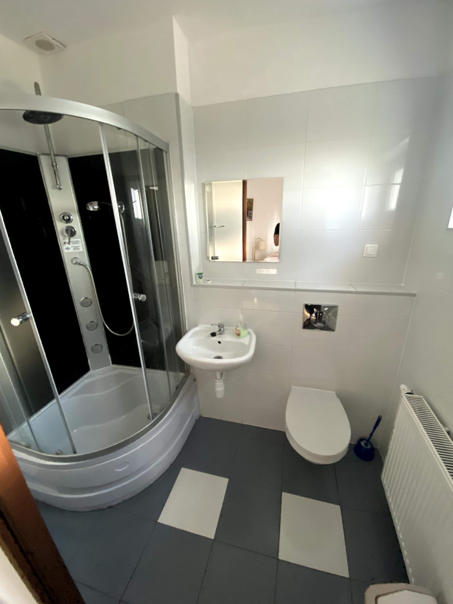 Wnętrze łazienki z kabiną prysznicową z czarnym panelem, umywalką na postumencie, toaletą i grzejnikiem żeberkowym, utrzymane w bieli i szarości.