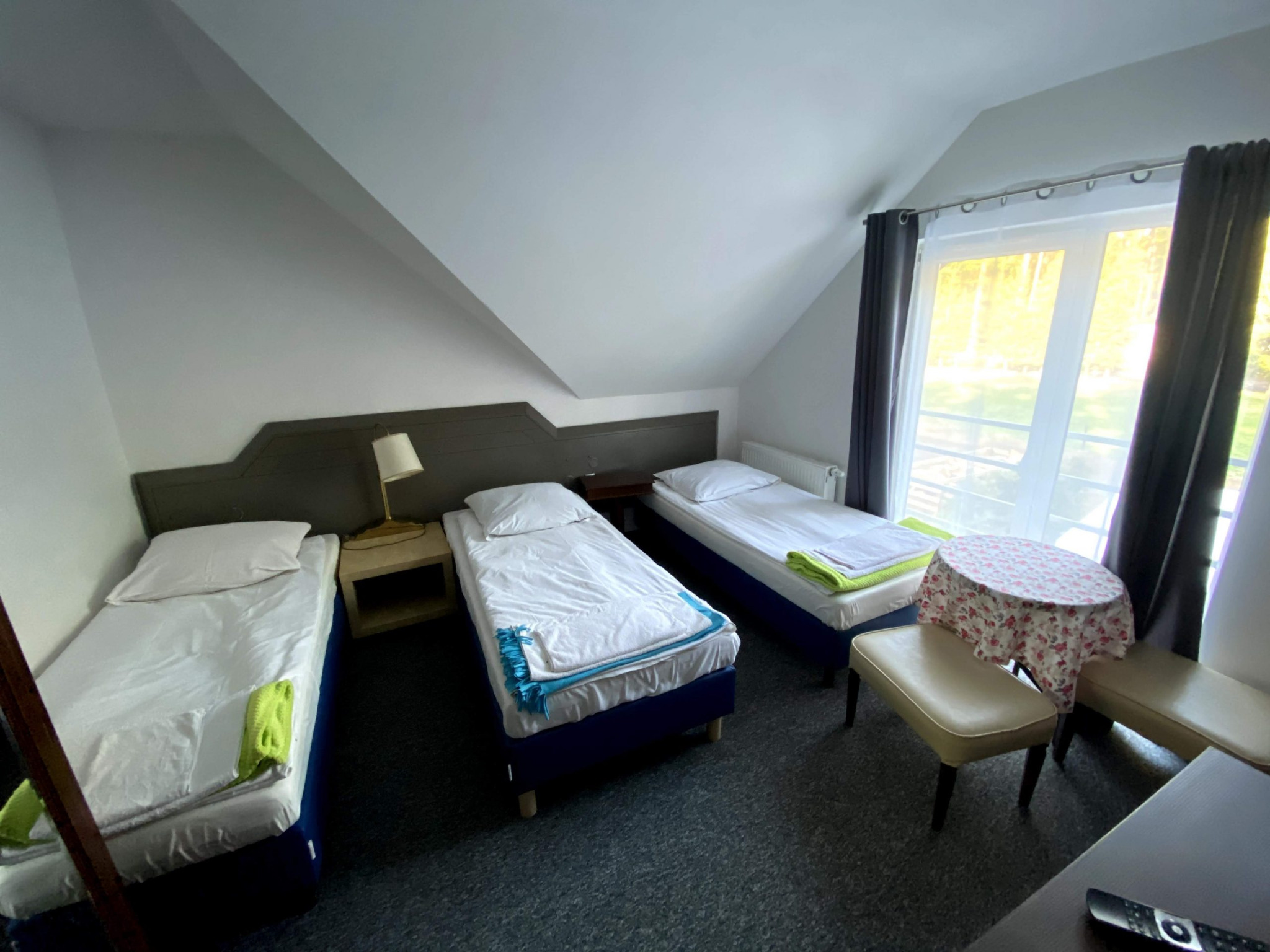 Pokój z trzema pojedynczymi łóżkami w pensjonacie w Poznaniu, nakryte białą pościelą i kolorowymi ręcznikami, stolik z kwiecistym obrusem i krzesła, okno z widokiem na zieleń.