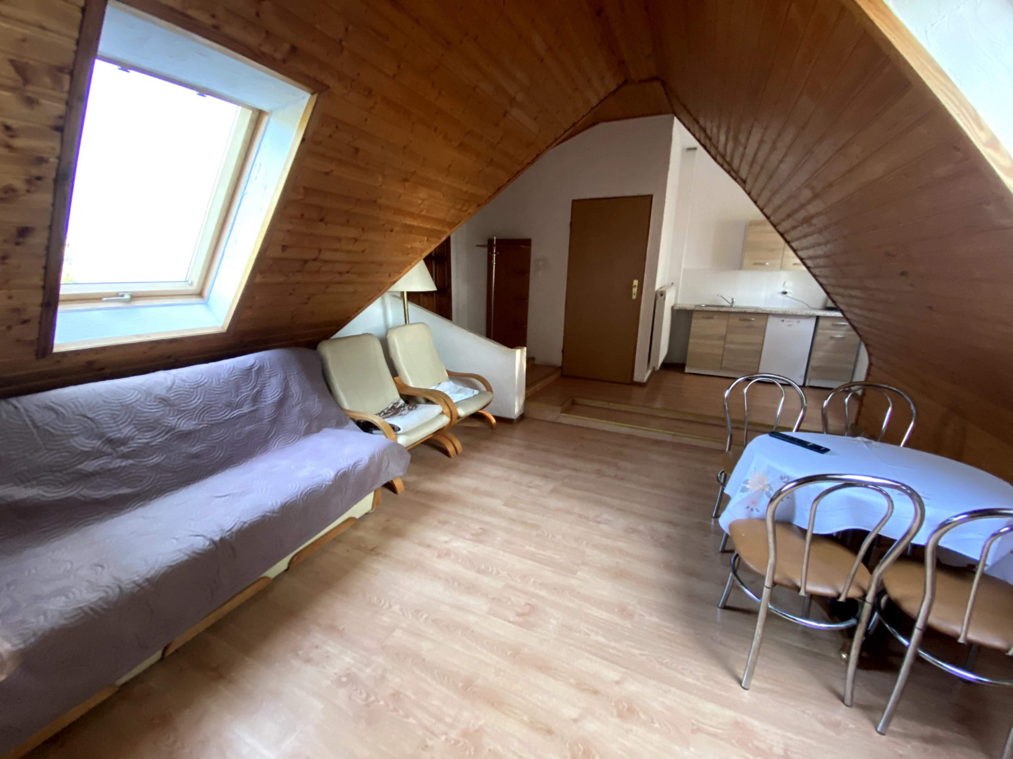 Pokój na poddaszu z drewnianym wykończeniem, widoczny aneks kuchenny, stół z krzesłami i rozkładana sofa. Na stole leży pilot do telewizora.