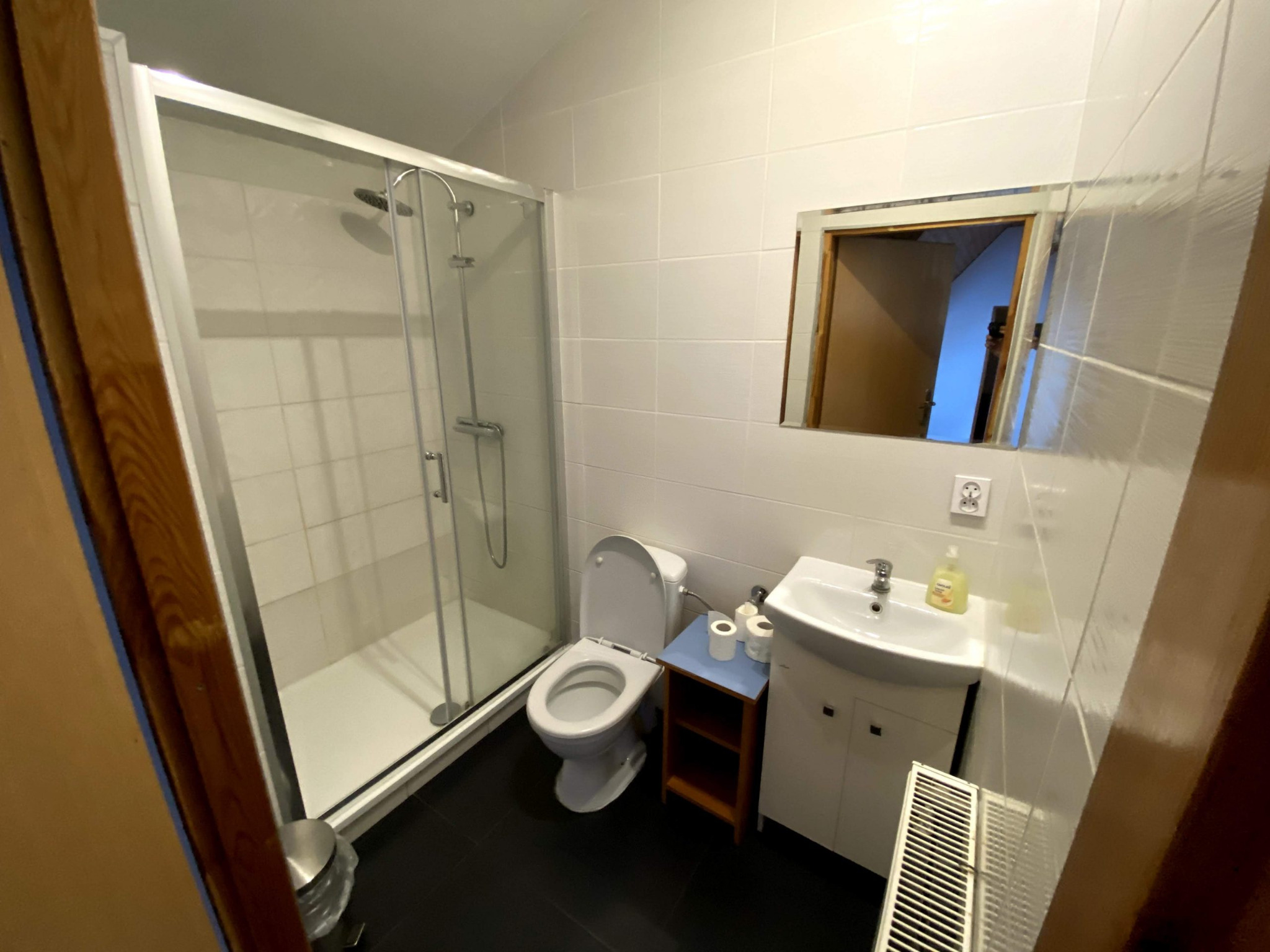 Wnętrze łazienki z kabiną prysznicową, toaletą, umywalką z szafką i grzejnikiem, wykończone białymi płytkami i czarną podłogą.