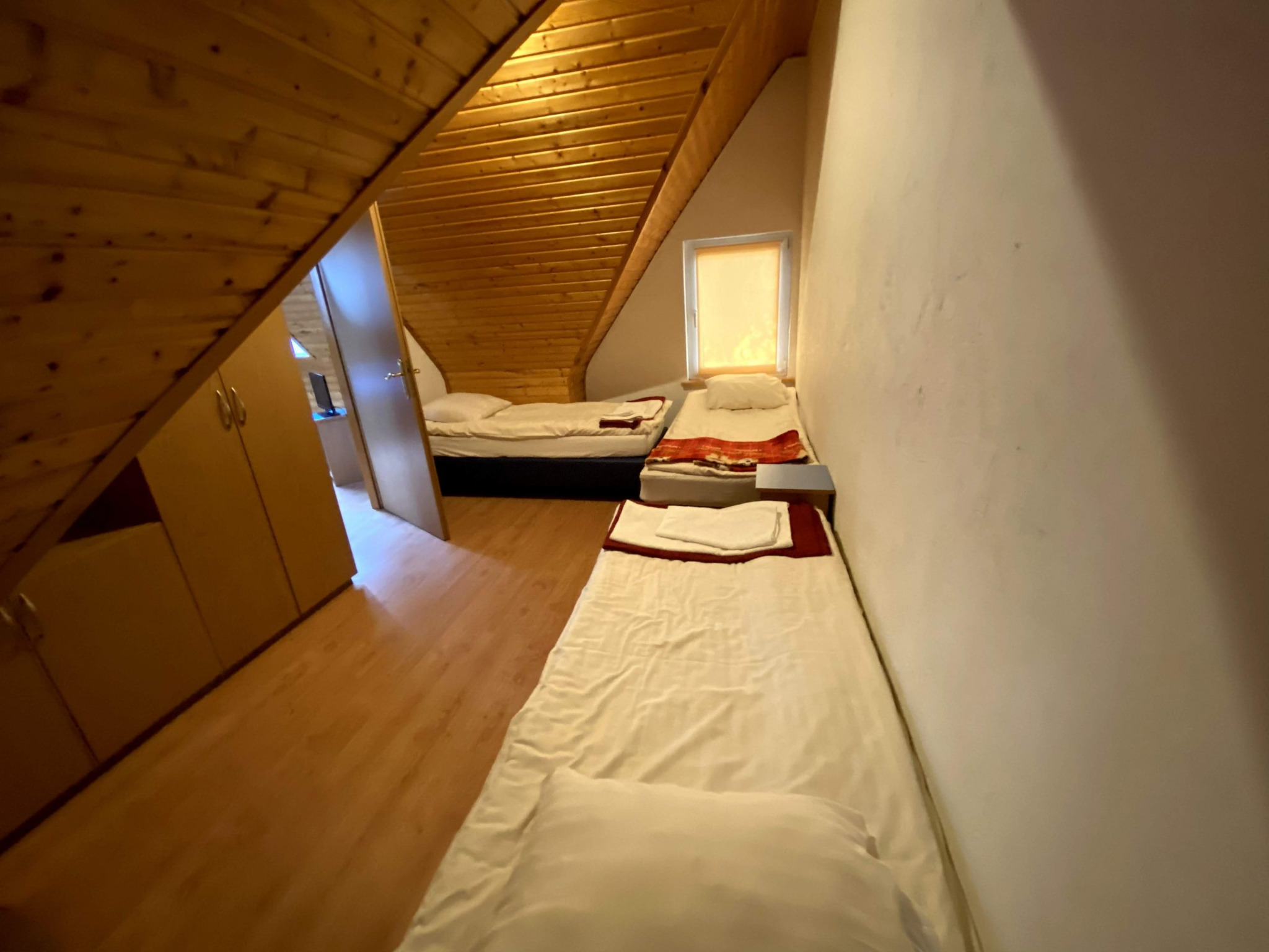 Pokój na poddaszu z trzema pojedynczymi łóżkami, drewnianą boazerią na suficie i szafą z jasnego drewna.