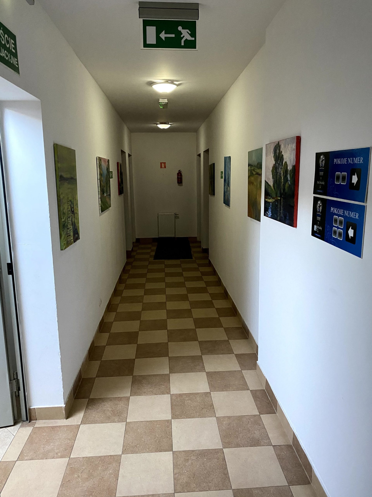 Korytarz w budynku z białymi ścianami, szachownicą na podłodze i obrazami na ścianach, tabliczki z numerami pokoi po prawej stronie, znak wyjścia ewakuacyjnego na suficie.