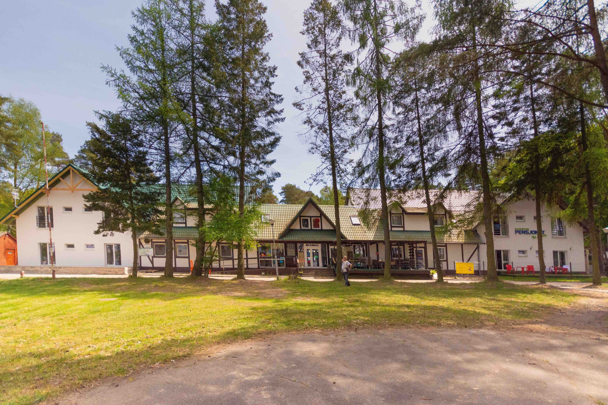 Budynek pensjonatu z zielonym dachem, otoczony wysokimi drzewami, z widocznym trawnikiem i osobą stojącą przed wejściem.