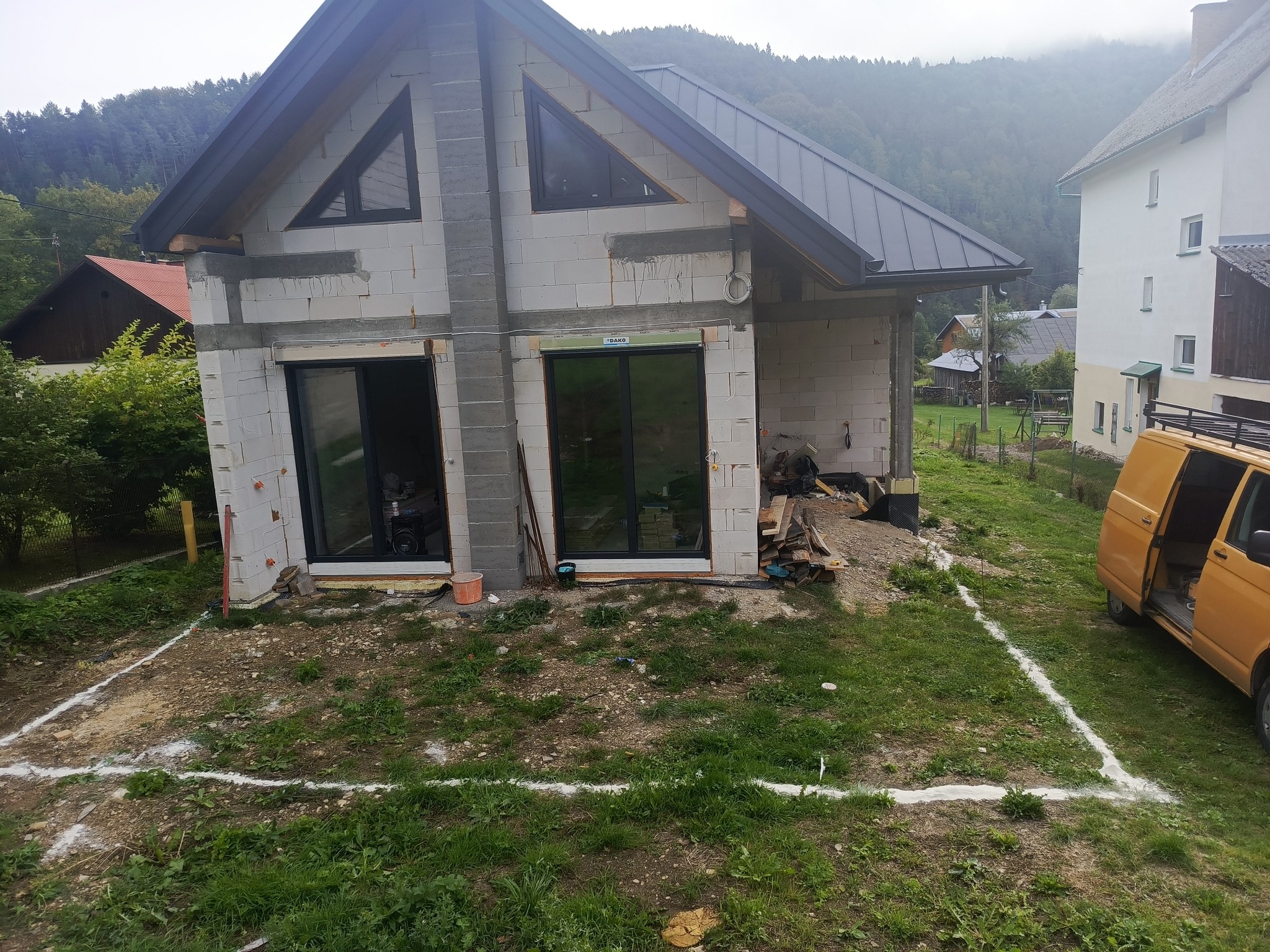 Budowa domu jednorodzinnego w stanie surowym otwartym: ściany z betonu komórkowego, czarne okna, widoczne fundamenty i zarys budynku na trawie. Górzysty krajobraz w tle.