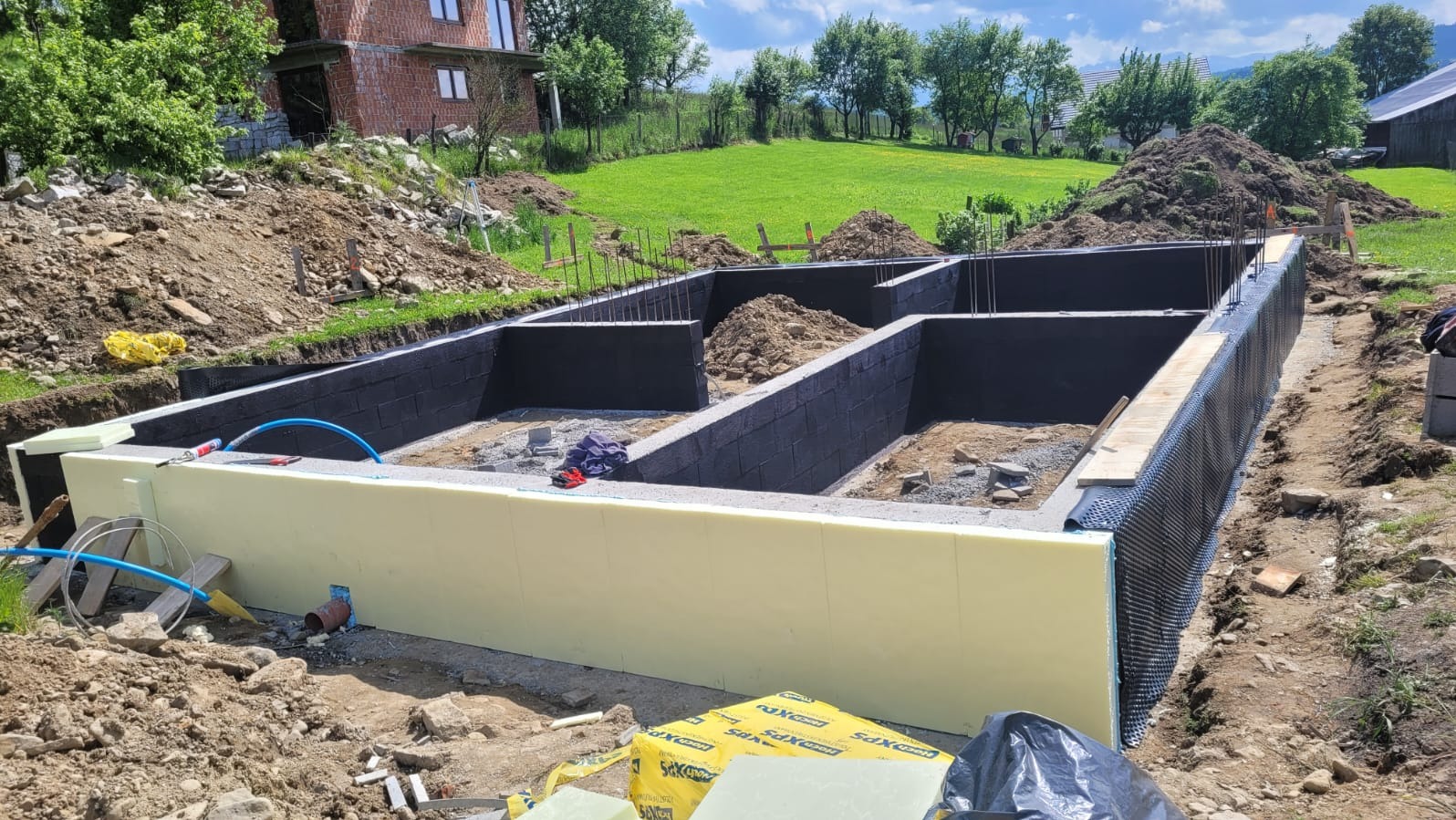 Fundamenty domu w trakcie budowy: ściany z bloczków pomalowane na czarno, ocieplone styropianem, z widocznym zbrojeniem i izolacją przeciwwilgociową. W tle zielony krajobraz.