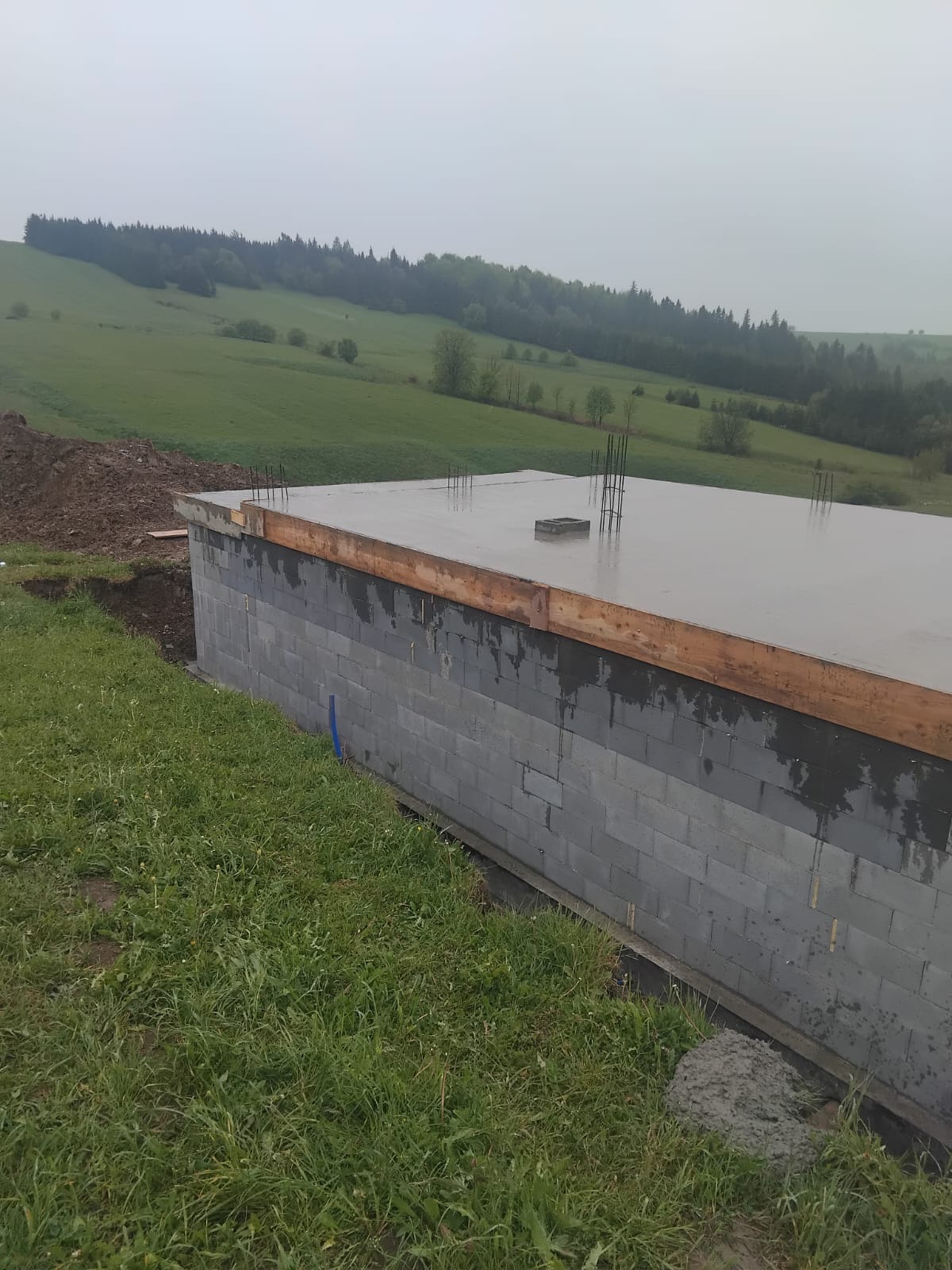 Ściana fundamentowa z bloczków betonowych, zalana betonem, z wystającymi prętami zbrojeniowymi, na tle zielonych wzgórz w pochmurny dzień.