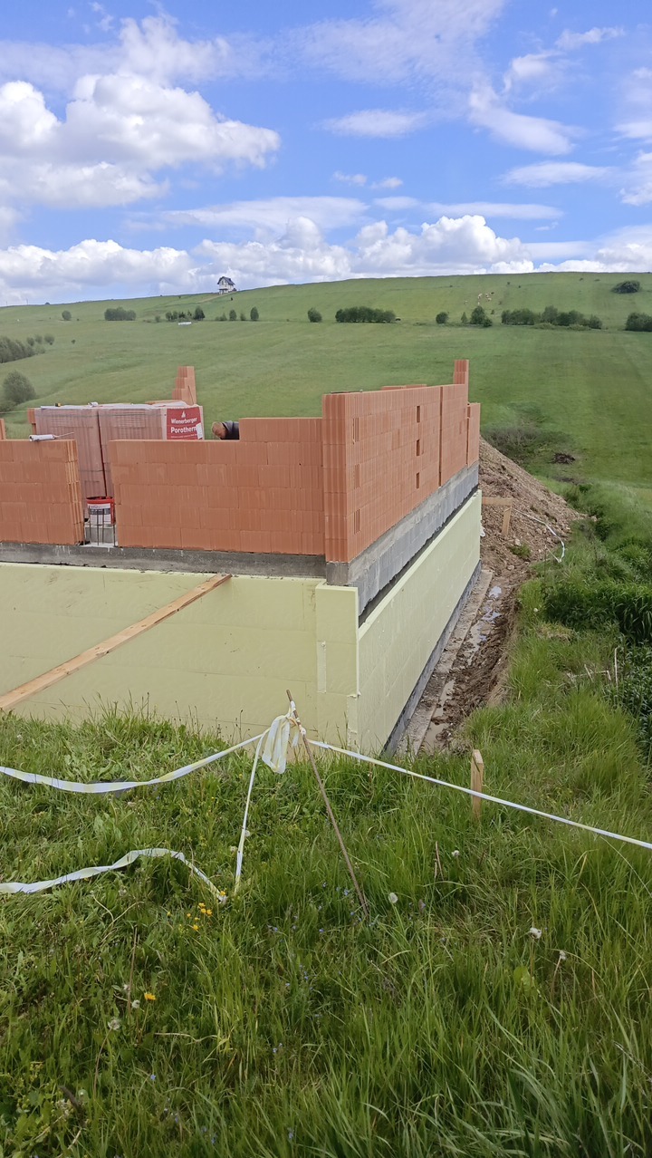 Wznoszenie ścian z czerwonej cegły Porotherm na fundamencie ocieplonym styropianem, na tle zielonego wzgórza i błękitnego nieba z chmurami.