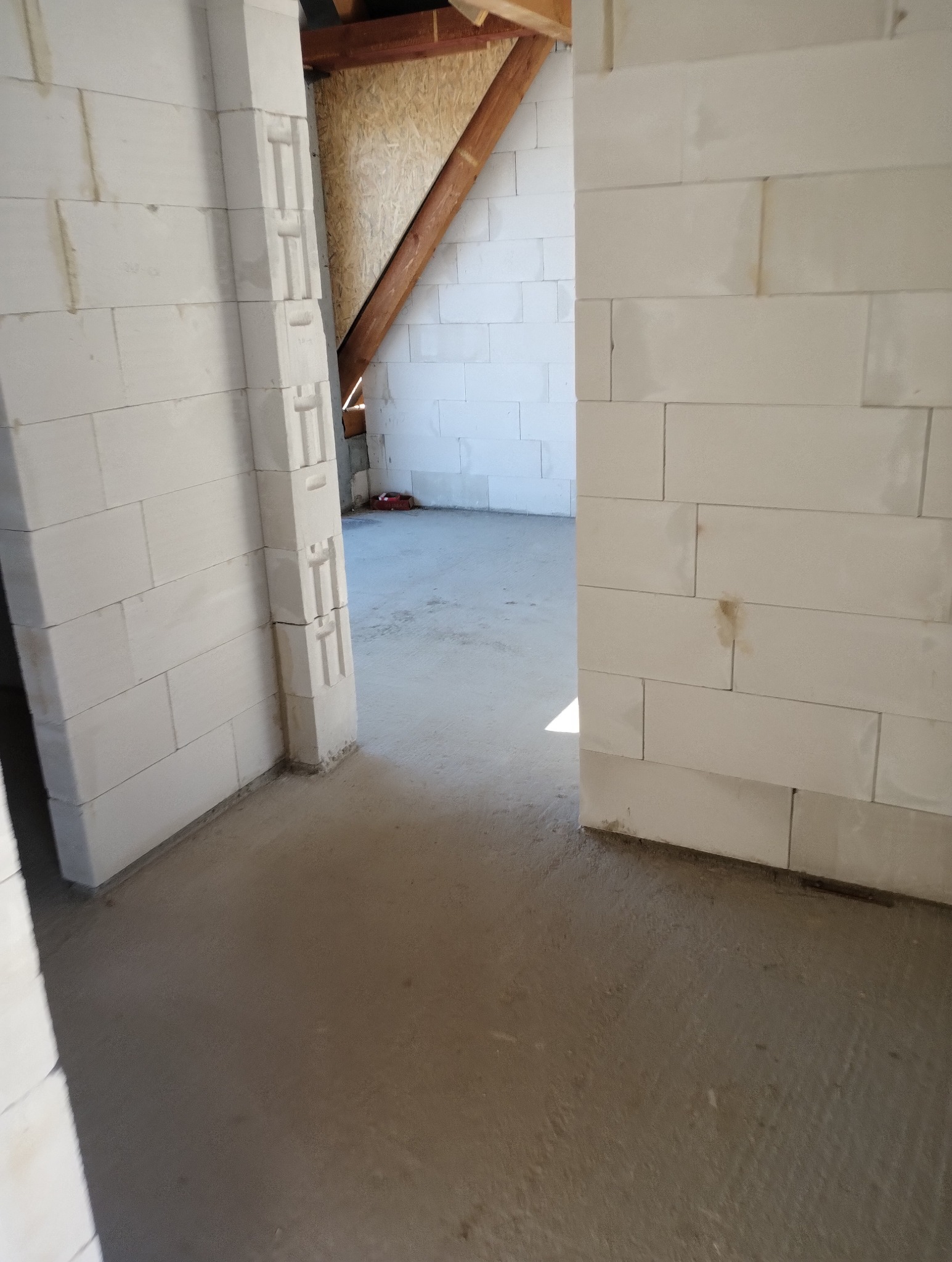 Wnętrze w trakcie budowy: ściany z białych bloczków, widoczna konstrukcja dachu z drewnianymi belkami i płytą OSB. Surowy beton na podłodze, widok na kolejne pomieszczenie.