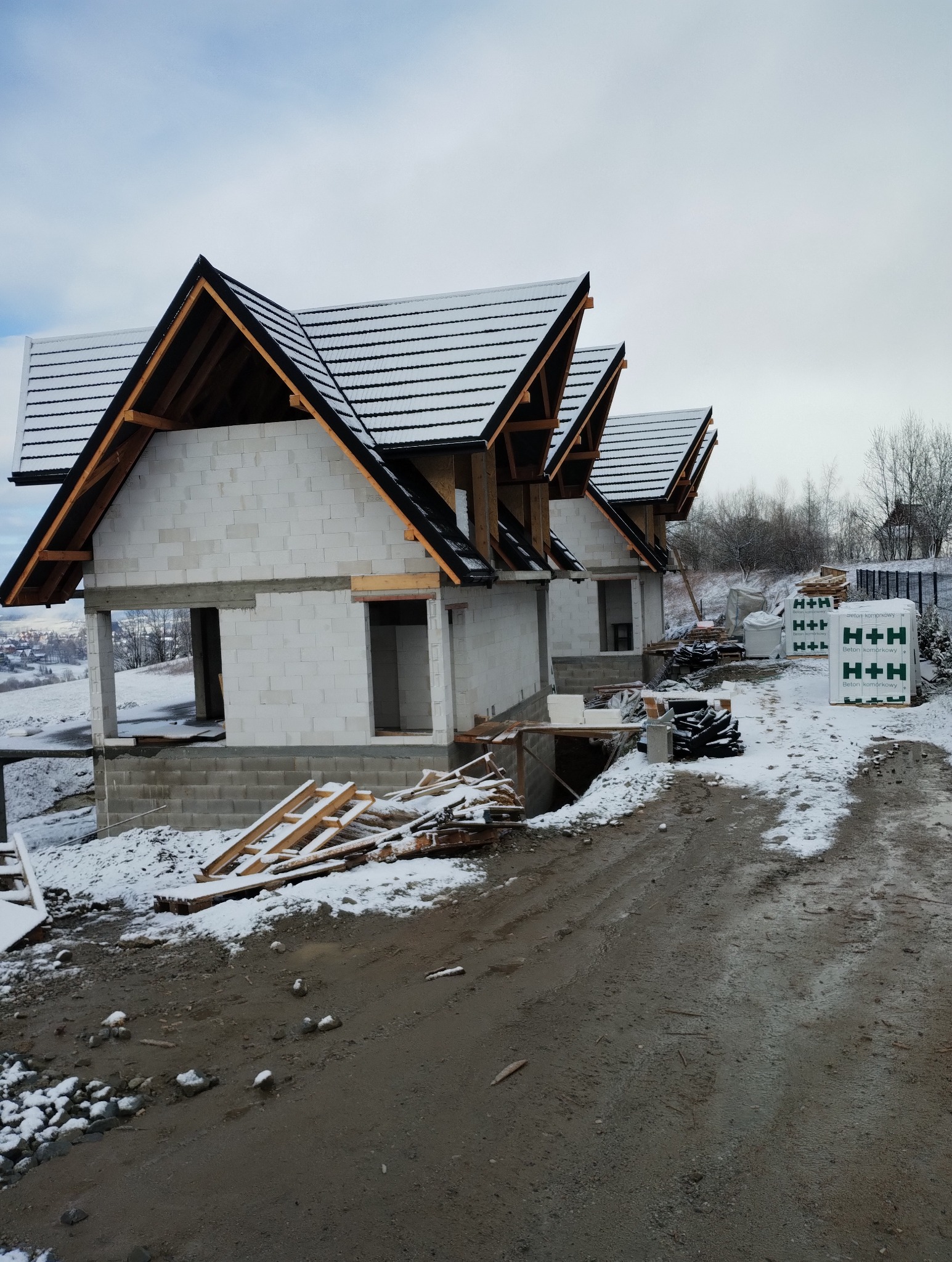 Murowanie ścian z betonu komórkowego H+H w zimowej scenerii. Dwa budynki w stanie surowym otwartym, z widoczną konstrukcją dachu i śniegiem na dachówce.