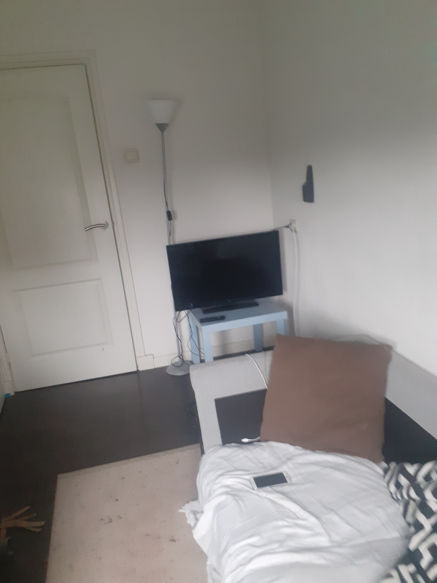 Pokój z ciemną podłogą, białą ścianą i drzwiami, lampą stojącą, telewizorem na niebieskim stoliku i częściowo widocznym łóżkiem z brązową poduszką i telefonem. Ujęcie z niskiej perspektywy.