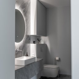 Knyf Studio - Nowoczesna łazienka w odcieniach szarości z marmurową teksturą, podświetlanym okrągłym lustrem, minimalistyczną umywalką nablatową i wiszącą toaletą.
