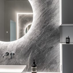 Knyf Studio - Minimalistyczna łazienka z umywalką nablatową, okrągłym lustrem z podświetleniem LED i marmurową ścianą w odcieniach szarości, uzupełniona o dozownik mydła Aesop i flakon perfum Jo Malone na półce.