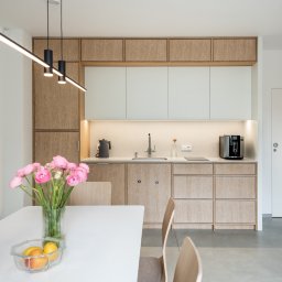 Knyf Studio - Nowoczesna kuchnia z drewnianymi i białymi szafkami, oświetleniem LED pod szafkami górnymi, czarnym ekspresem do kawy i czajnikiem na blacie, oraz wazonem z różowymi kwiatami na białym stole jadalnym.