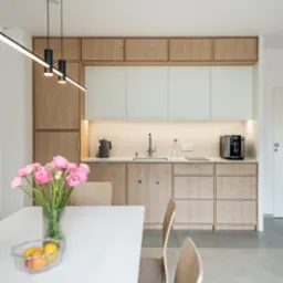 Nowoczesna kuchnia z drewnianymi i białymi szafkami, oświetleniem LED pod szafkami górnymi, czarnym ekspresem do kawy i czajnikiem na blacie, oraz wazonem z różowymi kwiatami na białym stole jadalnym.