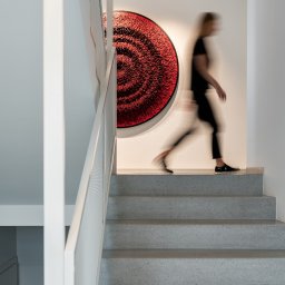 Knyf Studio - Betonowe schody z białą balustradą, czerwona okrągła dekoracja ścienna, rozmyta sylwetka kobiety schodzącej po schodach.