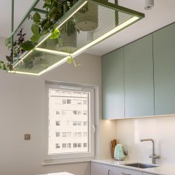Knyf Studio - Nowoczesna kuchnia z zielonymi szafkami i oryginalną lampą-konstrukcją z roślinami. Minimalistyczny design, jasne wnętrze, okno z widokiem na budynek.