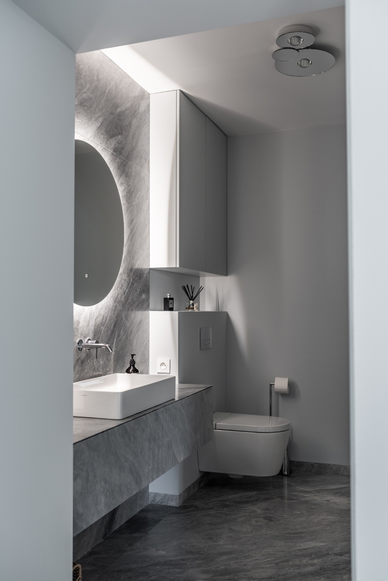 Nowoczesna łazienka w odcieniach szarości z marmurową teksturą, podświetlanym okrągłym lustrem, minimalistyczną umywalką nablatową i wiszącą toaletą.