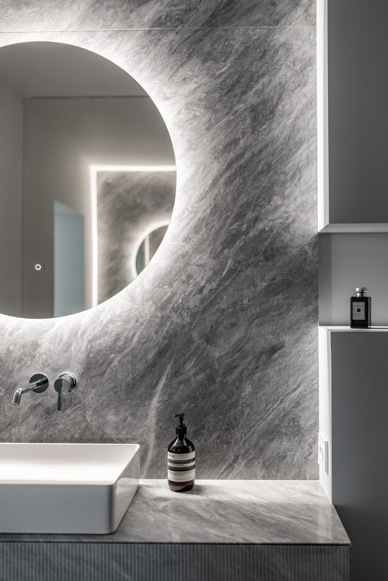 Minimalistyczna łazienka z umywalką nablatową, okrągłym lustrem z podświetleniem LED i marmurową ścianą w odcieniach szarości, uzupełniona o dozownik mydła Aesop i flakon perfum Jo Malone na półce.